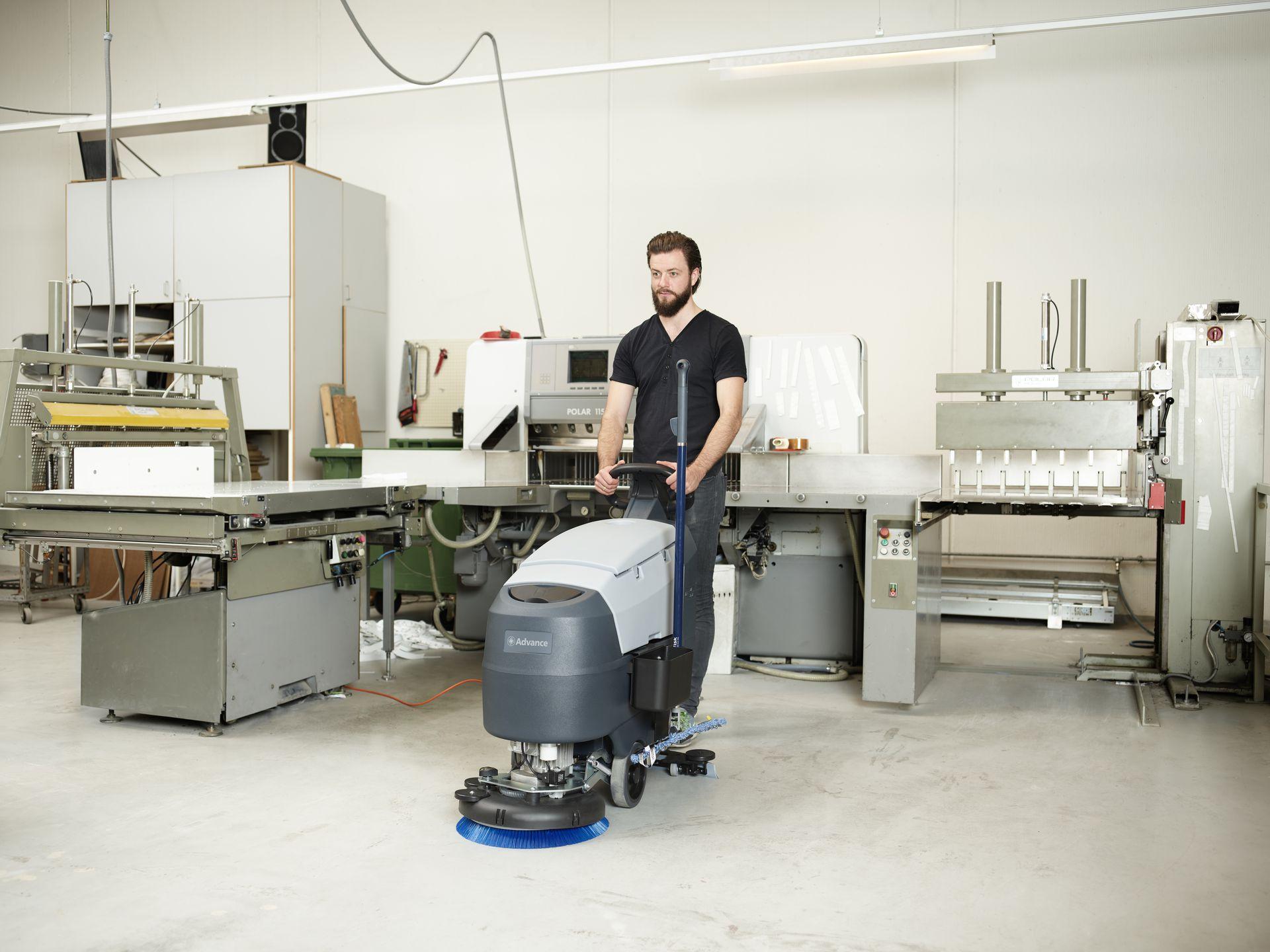 Worker operating a floor scrubber around machinery inside a manufacturing workshop.